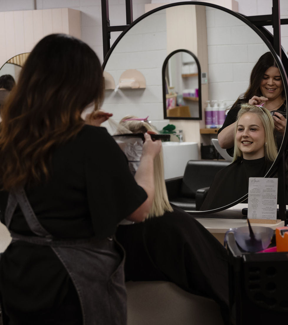 Hairdresser styling a client's hair in a salon setting with mirrors and salon equipment.